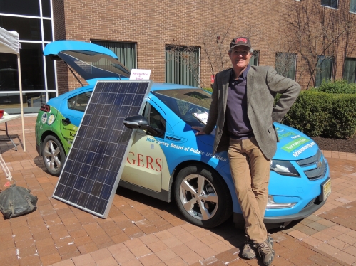 Male faculty wearing a baseball cap leaning against a solar car with the solar panel leaning on the side of the car with the back hatch open. 
