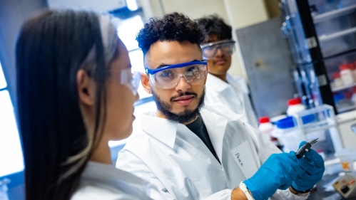 Two male students and one female student working in lab wearing safety goggles and a white lab coats..