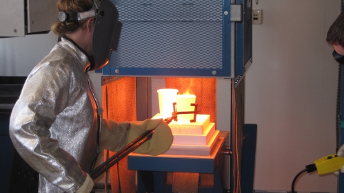 Woman wearing aluminized safety and jacket loading a crucible into a glass-melting furnace.