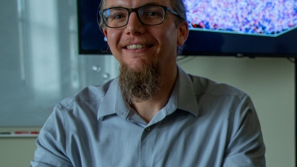 White male with glasses and a beard smiles at camera. He's wearing a light blue open necked shirt. Behind him is an abstract image on a monitor.
