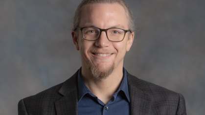 head shot of male with hair pulled into ponytail sporting a beard and glasses wearing a dark grey suit jacket and dark blue button down shirt