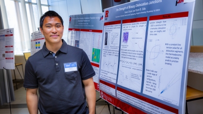 Asian male wearing a blue polo shirt and a name tag as he stands beside his poster presentation.