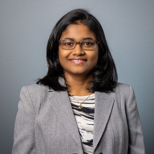 Head shot of Asian woman with shoulder length black hair and eyelgasses wearing a grey suit jacket and black and white patterned shirt.