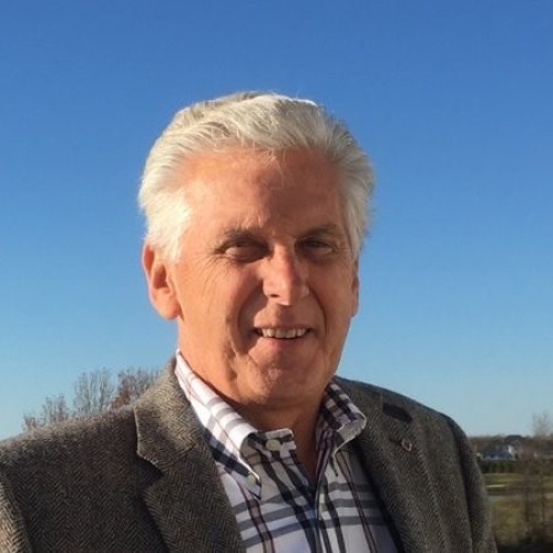 Outdoor head shot of elderly man with short white hair wearing a patterned shirt and bronw suit jacket.