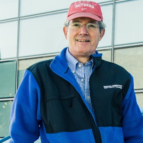 Headshot of male wearing eyeglasses, a red baseball cap, and a black and blue zip up jacket.