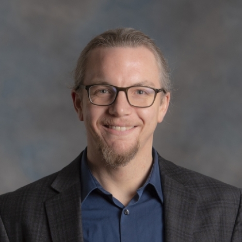 Headshot of male with facial hair and eyeglasses, wearing a dark suit and a dark bblue button down shirt.