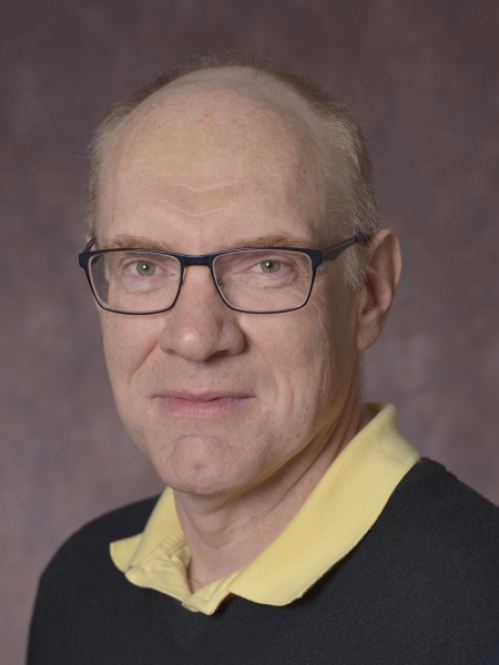Headshot of balding male wearing eyglasses, a black pullover sweater, and a yellow shirt.