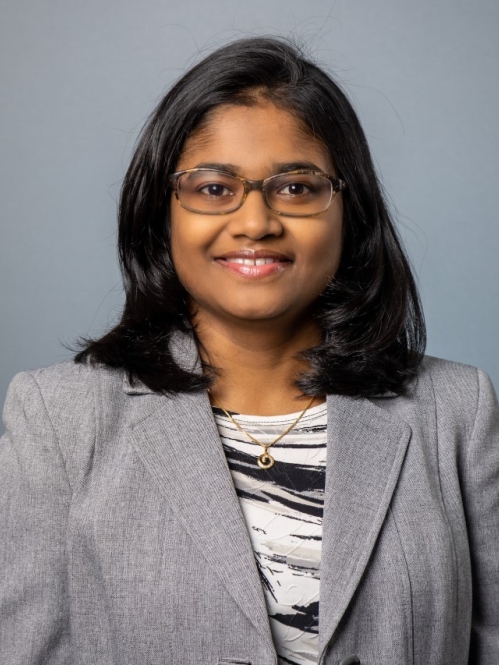 Head shot of Asian woman with shoulder length black hair and eyelgasses wearing a grey suit jacket and black and white patterned shirt.