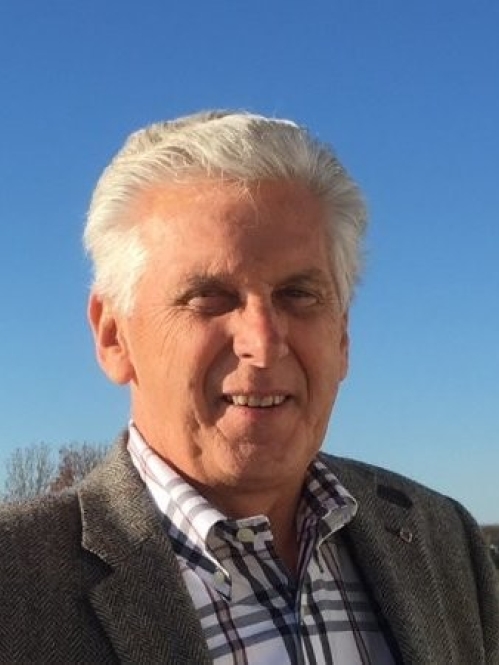 Outdoor head shot of elderly man with short white hair wearing a patterned shirt and bronw suit jacket.