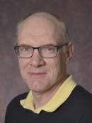 Headshot of balding male wearing eyglasses, a black pullover sweater, and a yellow shirt.