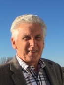 Outdoor head shot of elderly man with short white hair wearing a patterned shirt and bronw suit jacket.