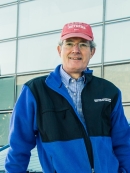 Headshot of male wearing eyeglasses, a red baseball cap, and a black and blue zip up jacket.