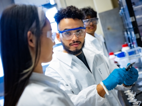 Two male students and one female student working in lab wearing safety goggles and a white lab coats..