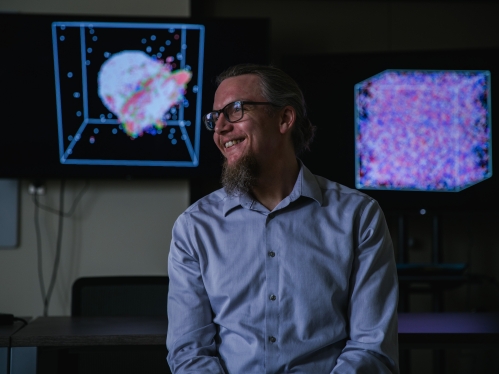 Head shot of male professor with beard and eyeglasses looking to his right with two large television screens behind him.