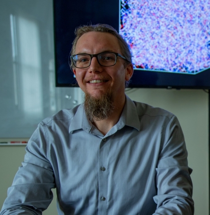 White male with glasses and a beard smiles at camera. He's wearing a light blue open necked shirt. Behind him is an abstract image on a monitor.