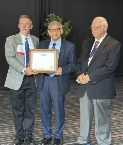 Award plaque presented to a man with white hair and eyeglasses flanked by two other men, all wearing suits and ties