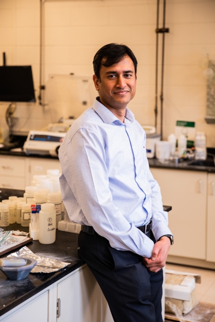 Professor leaning against a counter in a lab. He has brown hair and is wearing a light blue open collared shirt and dark pants.