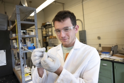 Male student working in laboratory wearing safety goggles, a white lab coat, and gloves.